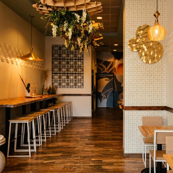 A restaurant interior with a long bar, stools, small tables, and decorative lighting.